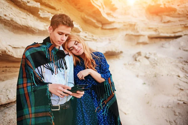 Beautiful woman and handsome man wrapped in a blanket. They are smiling and looking at the screen of a tablet on the background of a sand quarry.