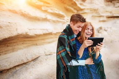 Güzel kadın ve yakışıklı bir battaniyeye sarılmış. Onlar gülümseyen ve bir tablet ekranında bir kum ocağı arka plan üzerinde seyir.