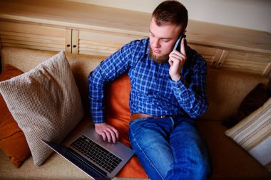 Onun smartphone kullanarak çevrimiçi bankacılık-sıçrama laptop ile kanepede oturan genç adam.