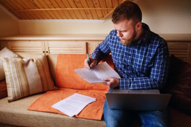 Genç adam eve bir dizüstü bilgisayarda çalışırken belgeleri tutan bir kanepede oturan eğitim.