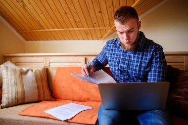 Genç adam eve bir dizüstü bilgisayarda çalışırken belgeleri tutan bir kanepede oturan eğitim.