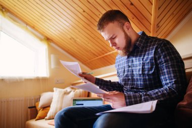 Genç adam eve bir dizüstü bilgisayarda çalışırken belgeleri tutan bir kanepede oturan eğitim.