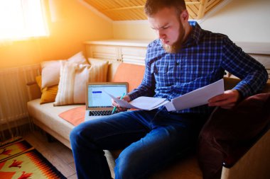 Genç adam eve bir dizüstü bilgisayarda çalışırken belgeleri tutan bir kanepede oturan eğitim.