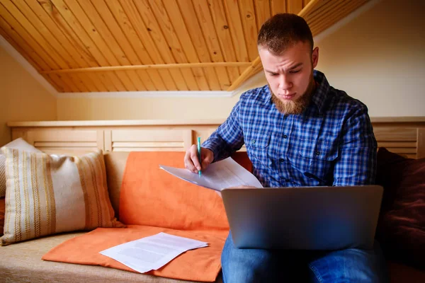Genç adam eve bir dizüstü bilgisayarda çalışırken belgeleri tutan bir kanepede oturan eğitim.