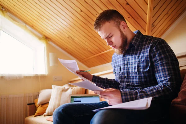 Genç adam eve bir dizüstü bilgisayarda çalışırken belgeleri tutan bir kanepede oturan eğitim.