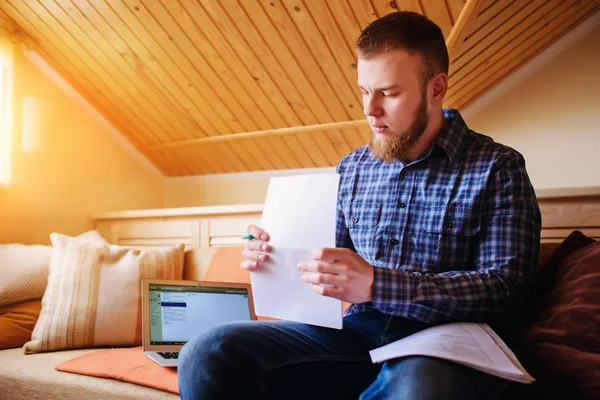 Genç adam eve bir dizüstü bilgisayarda çalışırken belgeleri tutan bir kanepede oturan eğitim.