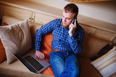 Onun smartphone kullanarak çevrimiçi bankacılık-sıçrama laptop ile kanepede oturan genç adam.