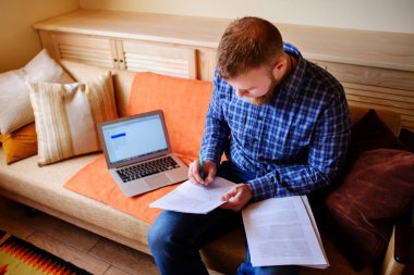 Genç adam eve bir dizüstü bilgisayarda çalışırken belgeleri tutan bir kanepede oturan eğitim.