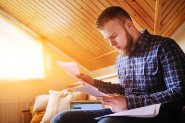 Genç adam eve bir dizüstü bilgisayarda çalışırken belgeleri tutan bir kanepede oturan eğitim.