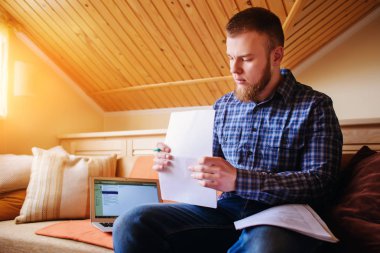 Genç adam eve bir dizüstü bilgisayarda çalışırken belgeleri tutan bir kanepede oturan eğitim.