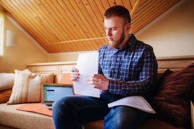 Genç adam eve bir dizüstü bilgisayarda çalışırken belgeleri tutan bir kanepede oturan eğitim.