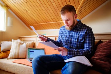 Genç adam eve bir dizüstü bilgisayarda çalışırken belgeleri tutan bir kanepede oturan eğitim.