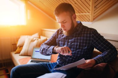 Genç adam eve bir dizüstü bilgisayarda çalışırken belgeleri tutan bir kanepede oturan eğitim.