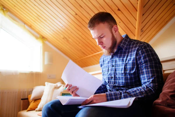 Young man studying from home sitting on a sofa holding documents while working on a laptop computer.