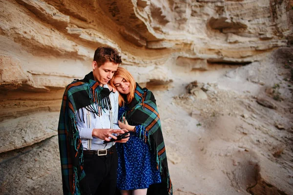 Beautiful woman and handsome man wrapped in a blanket. They are smiling and looking at the screen of a tablet on the background of a sand quarry.