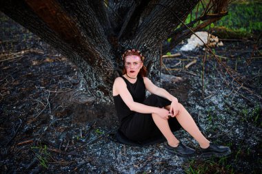 Menekşe Parlatıcı yüzünde güzel bir genç kadınla yanmış bir ağacın yanında duruyor.