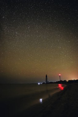 Siluetleri iki deniz feneri kumlu plaj ve okyanus yıldızlı gökyüzü arka planı.