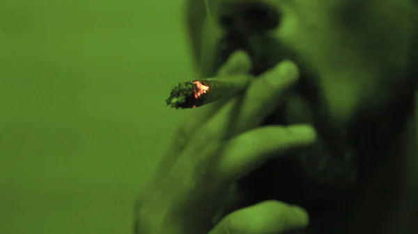 Um jovem fumando um jointer rolou com botões de erva dentro na luz verde close-up . — Fotografia de Stock