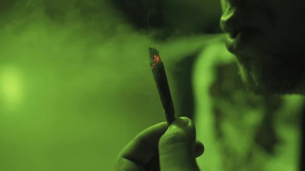 Młody facet palenia walcowane strugi z pąków chwastów wewnątrz w zbliżenie zielony światło. — Zdjęcie stockowe