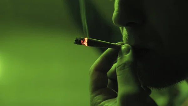 Un joven fumando una articulación enrollada con brotes de hierba en el interior en el primer plano de luz verde . —  Fotos de Stock