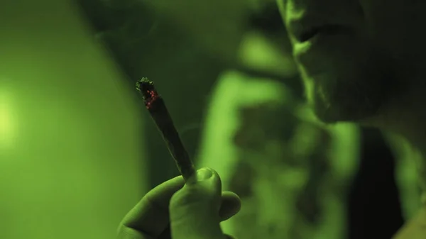 Ein junger Kerl, der im grünen Licht einen Rolljointer mit Unkrautknospen in Nahaufnahme raucht. — Stockfoto