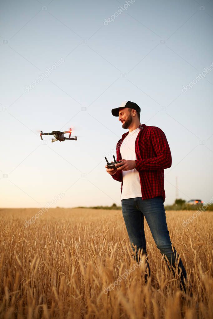 Farmer holds remote controller with his hands while quadcopter is flying on background. Drone hovers behind the agronomist in wheat field. Agricultural new technologies and innovations. Back view
