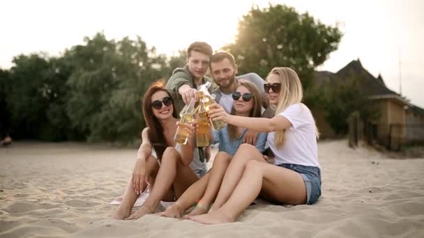 Eine Gruppe von Freunden genießt ein Getränk und entspannt sich am Strand bei Sonnenuntergang in Zeitlupe. junge Männer und Frauen trinken Bier sitzend auf einem Sand an einem warmen Sommerabend. — Stockvideo