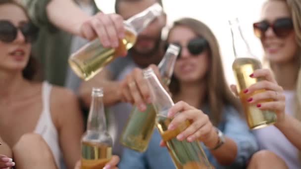 Groep vrienden plezier u geniet van een drankje en te ontspannen op het strand bij zonsondergang in slow motion. Jonge mannen en vrouwen drinken bier zittend op een zand in de warme zomeravond. — Stockvideo
