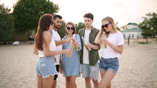 Grupo de amigos divirtiéndose disfrutando de una bebida y relajándose en la playa al atardecer en cámara lenta. Hombres y mujeres jóvenes beben cerveza de pie sobre una arena en la cálida noche de verano . — Vídeos de Stock