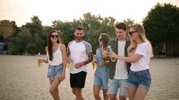 Grupo de amigos se divertindo desfrutando de uma bebida e relaxando na praia ao pôr do sol em câmera lenta. Os homens jovens e as mulheres bebem a cerveja que anda em uma areia na tarde quente do verão . — Vídeo de Stock