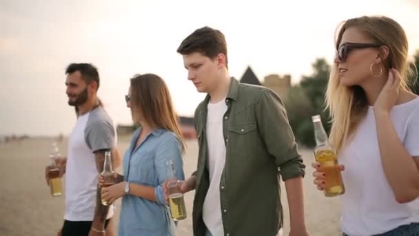 Ομάδα φίλων έχοντας διασκέδαση απολαμβάνοντας ένα ποτό και χαλάρωση στην παραλία το ηλιοβασίλεμα σε αργή κίνηση. Νεαροί άνδρες και γυναίκες να πίνουν μπίρα περπάτημα στην άμμο μια το ζεστό καλοκαιρινό βράδυ. — Αρχείο Βίντεο