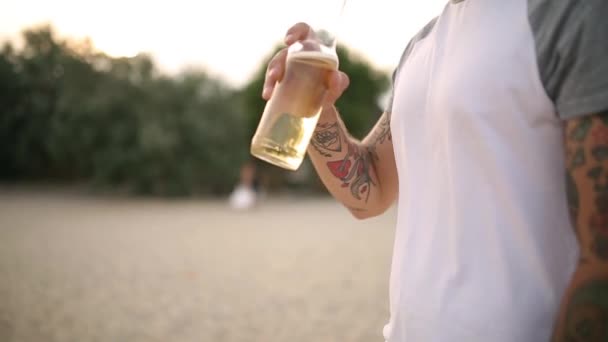 Jovem bonito tatuado barbudo caucasiano homem bebendo cerveja de garrafa de vidro na praia durante o pôr do sol, tiro steadycam, câmera lenta. Masculino sacia sede com bebida de limonada na costa do mar arenoso . — Vídeo de Stock