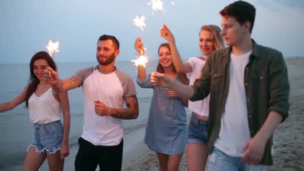 Amici che camminano, ballano e si divertono durante la festa notturna al mare con le luci scintillanti del bengala in mano. Giovani adolescenti che festeggiano sulla spiaggia con fuochi d'artificio. Scatto steadycam rallentato . — Video Stock