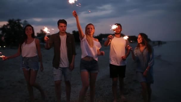 Amici che camminano, ballano e si divertono durante la festa notturna al mare con le luci scintillanti del bengala in mano. Giovani adolescenti che festeggiano sulla spiaggia con fuochi d'artificio. Scatto steadycam rallentato . — Video Stock