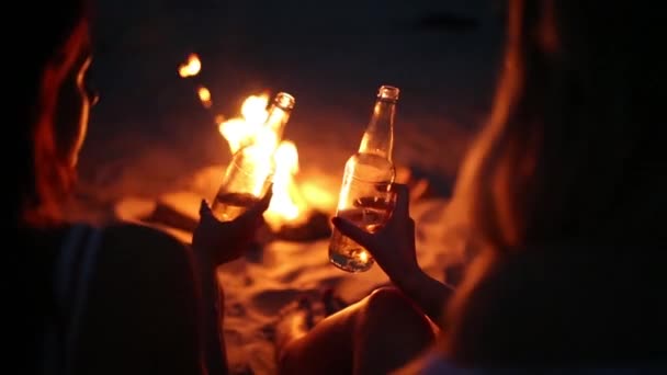 Fiesta en la playa al atardecer con hoguera. Amigos sentados alrededor de la hoguera, bebiendo cerveza y cantando a la guitarra. Hombres y mujeres jóvenes sostienen botellas de vidrio con bebidas singalong y animando . — Vídeos de Stock