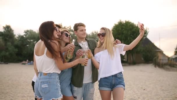 Grupp av vänner att ha kul njuter av en dryck och koppla av på stranden vid solnedgången i slow motion. Unga män och kvinnor dricka öl stående på en sand i den varma sommarkvällen. — Stockvideo