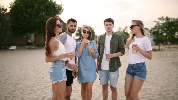 Skupina přátel baví užívat nápoj a odpočinek na pláži při západu slunce v pomalém pohybu. Mladí muži a ženy pijí pivo stojí na písku v teplé letní večery. — Stock video