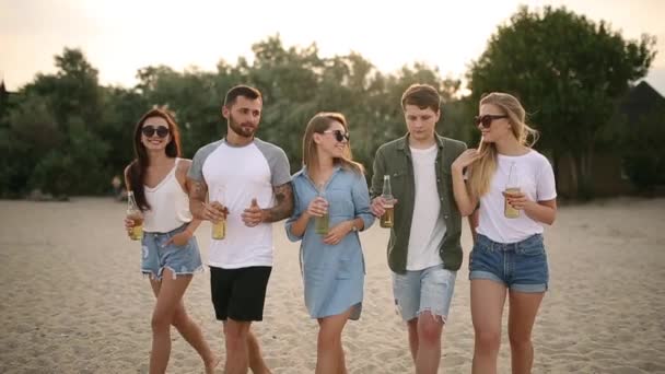 Group of friends having fun enjoying a beverage and relaxing on the beach at sunset in slow motion. Young men and women drink beer walking on a sand in the warm summer evening. — Stock Video