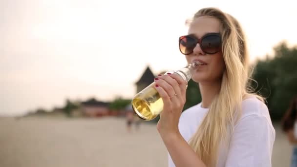 Jeune jolie femme marchant et buvant de la bière de bouteille en verre sur la plage pendant le coucher du soleil, tir steadycam, ralenti. Blonde dans les lunettes étanche la soif avec de la limonade boisson au bord de la mer sablonneuse . — Video