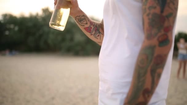 Joven guapo tatuado barbudo caucásico hombre beber cerveza de botella de vidrio en la playa durante la puesta del sol, steadycam disparo, cámara lenta. Macho sacia la sed con bebida de limonada en la orilla del mar arenoso . — Vídeo de stock
