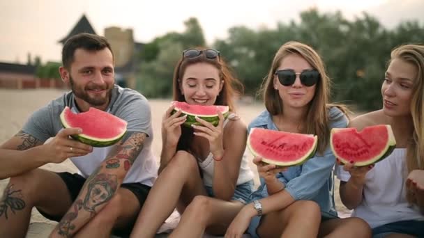 Happy vrienden eten watermeloen zittend op het zandstrand op vakantie. Jonge mannen en vrouwen het dragen van Blue Jeans Shorts. Vriendschap en begrip van de zomer — Stockvideo