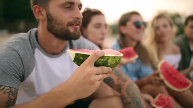 Mutlu arkadaş tatile Sandy Beach üzerinde oturan karpuz yeme. Kot şort giyen kadın ve erkekler. Dostluk ve yaz konsepti