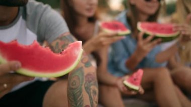 Mutlu arkadaş tatile Sandy Beach üzerinde oturan karpuz yeme. Kot şort giyen kadın ve erkekler. Dostluk ve yaz konsepti