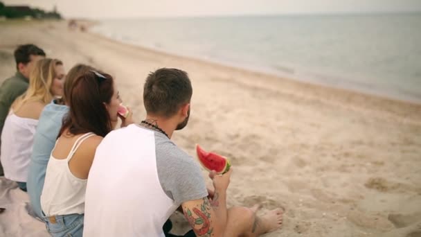 Happy Friends äta vattenmelon sitter på sandstranden på semester. Unga män och kvinnor som bär blå jeansshorts. Vänskap och sommaren koncept — Stockvideo