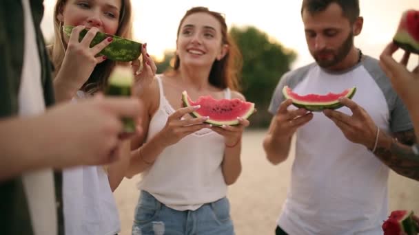 Happy Friends jedzenie arbuza stojący na piaszczysta plaża i rozmowy. Młodych kobiet i mężczyzn na sobie niebieskie dżinsy szorty w pobliżu morza na wakacjach. Przyjaźń i koncepcja lato — Wideo stockowe