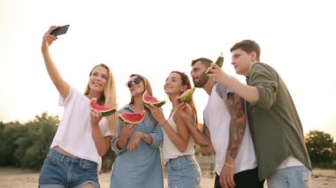 Sandy Beach ve alma Selfies Smartphone kamera ile ayakta karpuz yeme arkadaşlar. Genç erkek ve kadınların giydiği kot şort yakınındaki deniz ans Talking. Sağlıklı beslenme, Vegatarian gıda