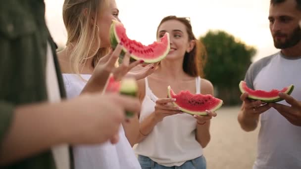 Felices amigos comiendo sandía de pie en Sandy Beach y charlando. Hombres y mujeres jóvenes con pantalones cortos vaqueros azules cerca del mar de vacaciones. Concepto de amistad y verano — Vídeo de stock