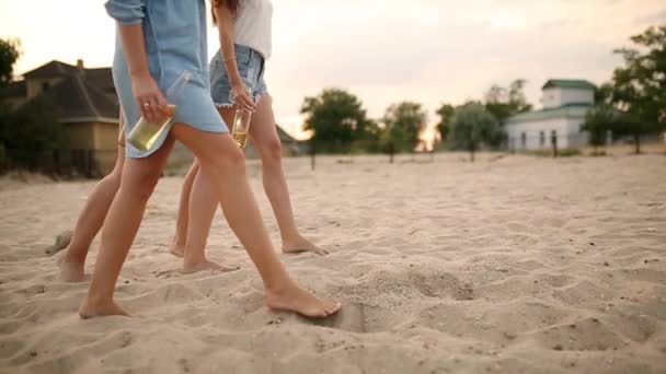 Vista aislada de tres pares de piernas delgadas femeninas con botellas de vidrio de bebida en manos que pisan la playa junto al mar al atardecer en cámara lenta. Las mujeres jóvenes beben cerveza caminando sobre una arena. Sin cara. . — Vídeos de Stock