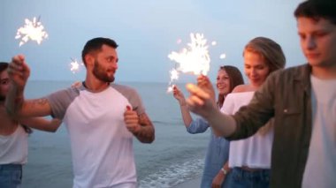 Arkadaşlar, yürüyüş bengal maytap ışıklar ellerinde ile deniz kenarında dans ve having fun gece boyunca parti. Havai fişek ile sahilde parti gençler. Ağır çekim steadycam vurdu.