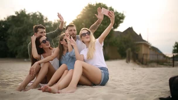 Tiro de um grupo de jovens amigos tirando uma selfie na praia. Homens e mulheres tirando fotos sentados em uma areia na noite quente de verão . — Vídeo de Stock